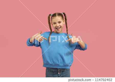 Cheerful young girl with pigtails in a blue sweater confidently pointing at herself against a pink background 114702402