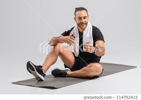 A man sits on a black fitness mat, holding a water bottle, catching his breath after a workout session. Wearing athletic shorts, a sleeveless top, and sports shoes, using smartphone A man sits on a black fitness mat, holding a water bottle, catching his breath after a workout session. Wearing athletic shorts, a sleeveless top, and sports shoes, using smartphone 114702415