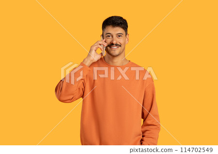 A man wearing an orange sweater is engrossed in a phone conversation. He holds a cell phone to his ear and gestures animatedly as he talks. 114702549