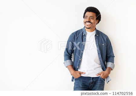A cheerful african american guy stands with hands in pockets against a white background, depicting a relaxed man at home A cheerful african american guy stands with hands in pockets against a white background, depicting a relaxed man at home 114702556