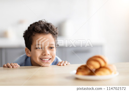 A cheerful african american boy peers at a plate of croissants, highlighting childhood curiosity and the anticipation of a tasty treat in a kitchen A cheerful african american boy peers at a plate of croissants, highlighting childhood curiosity and the anticipation of a tasty treat in a kitchen 114702572