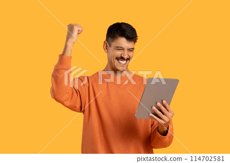 A man wearing an orange sweater is seen holding a tablet in his hands. He appears focused on the screen, engaged in tablet activities. 114702581