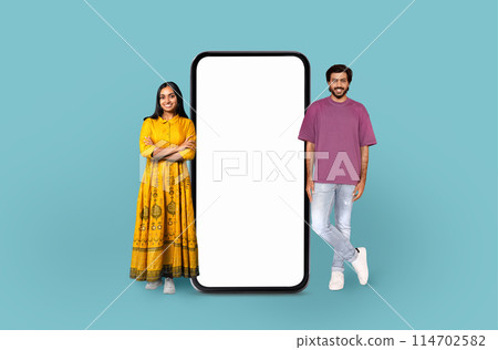 Indian woman in traditional dress and a man in casual wear stand beside a large blank smartphone screen, implying technology use 114702582