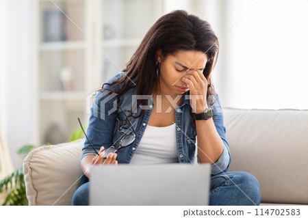 Overworked African American woman is seated on a couch, working on a laptop, removed her eyeglasses and rubbing her eyes, feeling exhausted 114702583