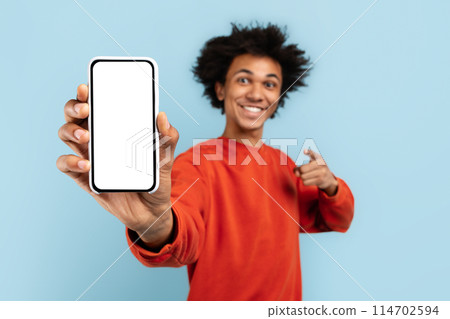 Cheerful black guy in an orange pullover promotes a blank smartphone screen, suitable for content display, blue isolated background Cheerful black guy in an orange pullover promotes a blank smartphone screen, suitable for content display, blue isolated background 114702594