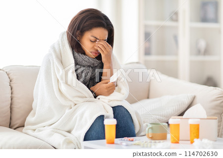 African American woman is seated on a couch, holding her hand to her face in a contemplative manner, have fever, suffering from cold 114702630