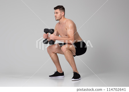 A man is squatting down while gripping two dumbbells in each hand. He is dressed in workout attire and appears focused on his exercise routine. 114702641