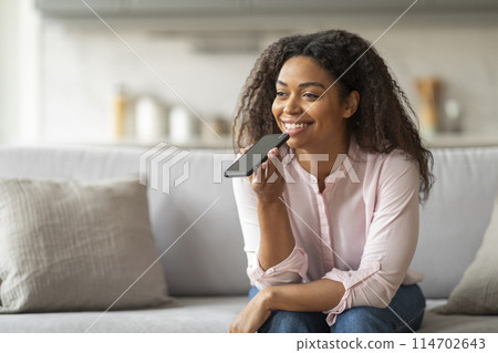 A cheerful young African American woman comfortably engages with her smartphone using voice command while relaxing on the sofa at home A cheerful young African American woman comfortably engages with her smartphone using voice command while relaxing on the sofa at home 114702643