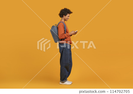 A modern black man in casual wear with backpack using smartphone, texting friends, isolated on orange backdrop A modern black man in casual wear with backpack using smartphone, texting friends, isolated on orange backdrop 114702705