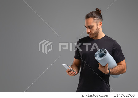 A man standing and holding a yoga mat in one hand and a cell phone in the other, ready for a workout or yoga session, possibly checking his phone for instructions or tracking his progress, copy space A man standing and holding a yoga mat in one hand and a cell phone in the other, ready for a workout or yoga session, possibly checking his phone for instructions or tracking his progress, copy space 114702706