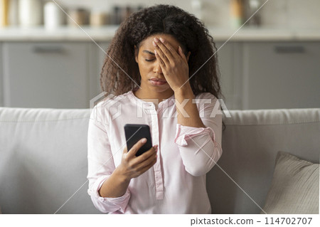 A photo capturing the tension of a young African American woman sitting on her couch, visibly overwhelmed as she looks at her smartphone in a neatly arranged living room 114702707