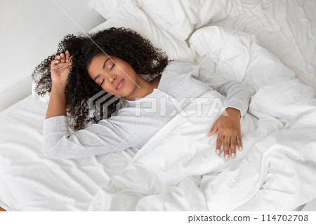 A woman is laying down in a bed with clean white sheets. She is wearing a comfortable outfit and appears to be relaxing or resting peacefully. 114702708