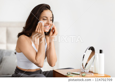 Smiling young woman in casual wear using a cotton pad to cleanse her face, sitting at a dressing table with cosmetics 114702709