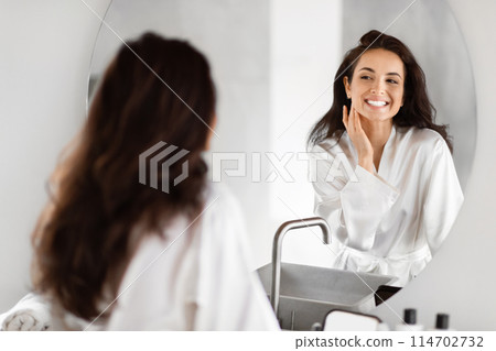 A woman looks at her reflection, showing the joy of a millennial's skin care ritual in a modern bathroom environment 114702732