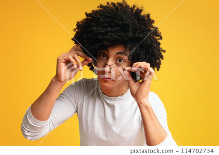 Kinda doubtful. Black millennial guy looking skeptically, lowing his glasses, orange studio background Kinda doubtful. Black millennial guy looking skeptically, lowing his glasses, orange studio background 114702734