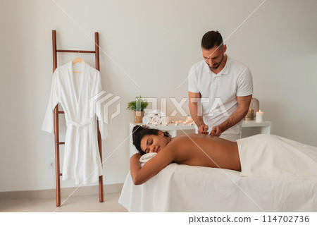 A man attentively lights candles near a serene African American woman at a spa, symbolizing the tranquil atmosphere created for people 114702736