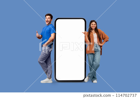 Young man and woman pose beside a large smartphone with white screen mockup copy space on a blue background 114702737
