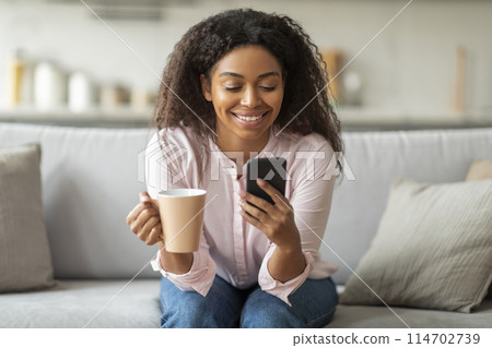 A relaxed afternoon scene where African American woman enjoys her leisure time by engaging with her mobile device, sitting comfortably on her living room sofa, drinking coffee 114702739