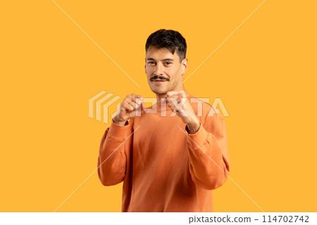 A confident young man raises his fists in a defensive stance, wearing a casual orange sweatshirt. He stands against a solid yellow backdrop 114702742