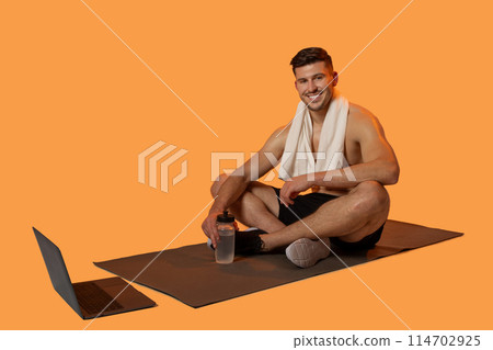 A cheerful man sits on a yoga mat with a white towel draped over his shoulders, holding a water bottle. With a laptop beside him, it appears he following an online workout or yoga session 114702925