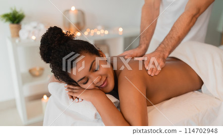 A close-up image of a black lady enjoying a soothing shoulder massage at a spa, with focus on the hands of the therapist and a relaxing atmosphere 114702948