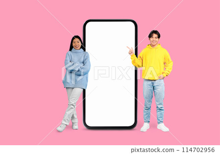 Happy Asian couple smiling warmly and gesturing towards a blank smartphone screen on a pink background 114702956