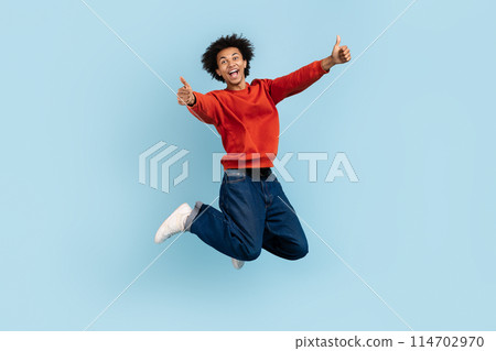 The image captures an african american zoomer guy mid-leap, thumbs up with an ecstatic expression, showcasing high spirits and freedom, against a blue isolated setting 114702970