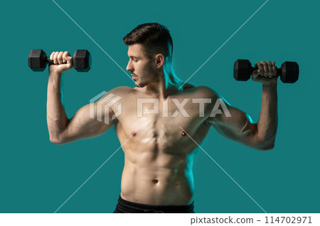 A shirtless man is standing while holding two dumbbells in his hands. He appears to be working out his muscles and strengthening his arms in a fitness setting. 114702971