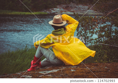 A tourist in a raincoat, hat and boots sits on the rocks next to the sea and looks into the distance. 114702993