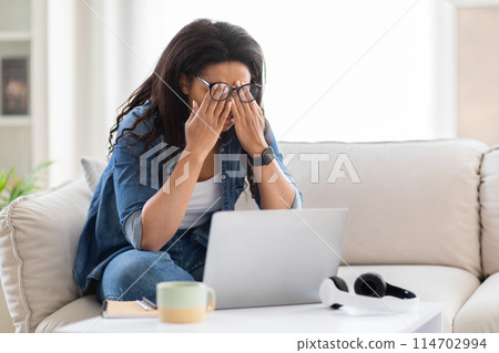 African American woman is shown sitting on a couch, her hands covering her eyes. She appears to be shielding her eyes from something, possibly light or a disturbing sight. 114702994