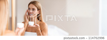 Facial skin treatment concept. Middle aged woman applying moisturizing cream on face standing wrapped in towel in front of mirror in bathroom, panorama with copy space 114702996