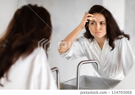 A woman in a white robe appears concerned as she examines her forehead for wrinkles in the bathroom mirror, embodying beauty anxieties 114703008