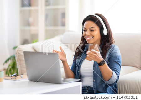 Smiling black woman is sitting on a beige couch with a laptop open in front of her, using headset, appears to be working or browsing the internet. 114703011