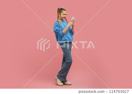 A cheerful young girl with braided hair is standing, happily looking at her smartphone against a pink background 114703027
