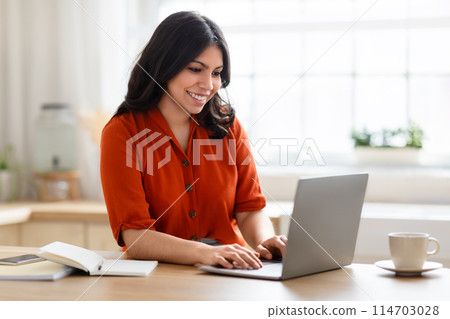 Happy middle eastern woman in an orange top smiling while typing on her laptop at a bright kitchen setting Happy middle eastern woman in an orange top smiling while typing on her laptop at a bright kitchen setting 114703028