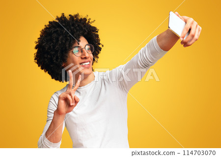 Young black guy making selfie with peace gesture on cellphone, orange studio background 114703070