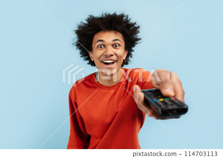 This image captures the excitement of a young black guy as he holds a TV remote control, representing leisure activities of a zoomer, isolated blue background 114703113