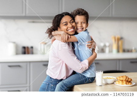 A young african american boy wraps his arms around his mother in a modern kitchen, illustrating the warmth and closeness within a black family 114703119