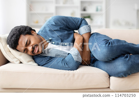 Unhealthy young black man lying on couch at home, touching his tummy, suffering from abdominal pain Unhealthy young black man lying on couch at home, touching his tummy, suffering from abdominal pain 114703250