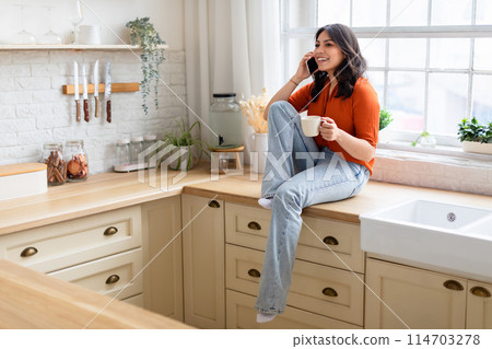 Cheerful middle eastern woman holds a coffee cup while having a phone conversation in her cozy kitchen setting Cheerful middle eastern woman holds a coffee cup while having a phone conversation in her cozy kitchen setting 114703278