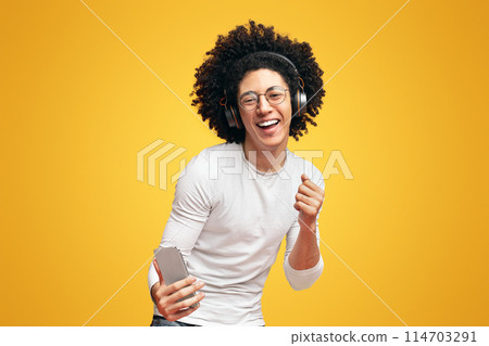 Handsome young african-american guy in wireless headphones listening to music online on smartphone and dancing, orange background 114703291