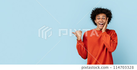 African american guy in orange sweater looking surprised and pointing sideways, isolated blue background, copy space 114703328