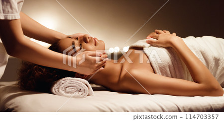 A serene African American woman lies on a spa bed receiving a gentle facial massage highlighting the concept of self-care and beauty 114703357
