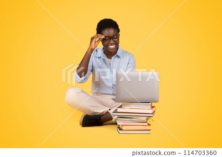 Engrossed African American woman wearing glasses, working on a laptop with a pile of books beside her, conveys focus and dedication, isolated on yellow 114703360