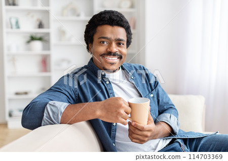 Cheerful african american guy sitting at home with a coffee mug, evoking warmth and morning routine Cheerful african american guy sitting at home with a coffee mug, evoking warmth and morning routine 114703369