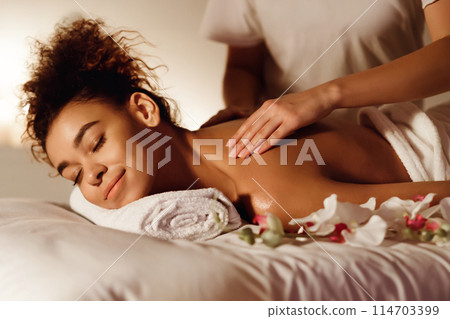 An African American woman smiles with pleasure while receiving a shoulder massage, suggesting a theme of blissful relaxation 114703399
