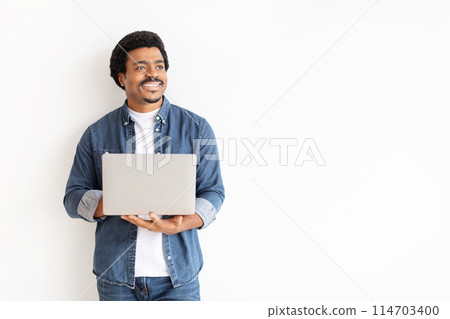 A cheerful man in casual denim attire holding a laptop computer and looking to the side with a hopeful expression, copy space 114703400