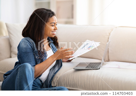 African American woman is seated on a couch, engrossed in reading a paper, focused on the content, holding the paper up with both hands. The room is well-lit, and she appears relaxed and attentive. 114703403