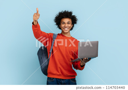This image features a young african american guy student wearing a sweater, with a laptop, giving a thumbs up against a blue isolated background This image features a young african american guy student wearing a sweater, with a laptop, giving a thumbs up against a blue isolated background 114703440