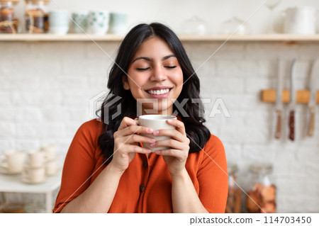 A content middle eastern woman in an orange blouse holds a white cup, eyes closed, with a kitchen setting in the background 114703450
